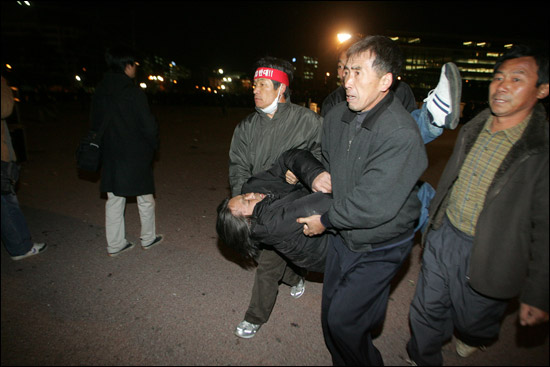 지난 15일 경찰에 폭행당해 쓰러진 고 전용철 농민을 다른 농민들이 발견해 들다시피 부축하고 공원을 빠져나가고 있다. 빨간 머리띠를 맨 이가 김장택 제주도연맹 조천읍지회장, 고인의 오른쪽 팔을 붙든 이가 정태문 성산읍지회장이다. [출처] <민중의소리>(voiceofpeople.org)