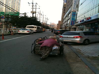 [설명] 폐지를 담은 손수레를 노인이 밀고 있다.